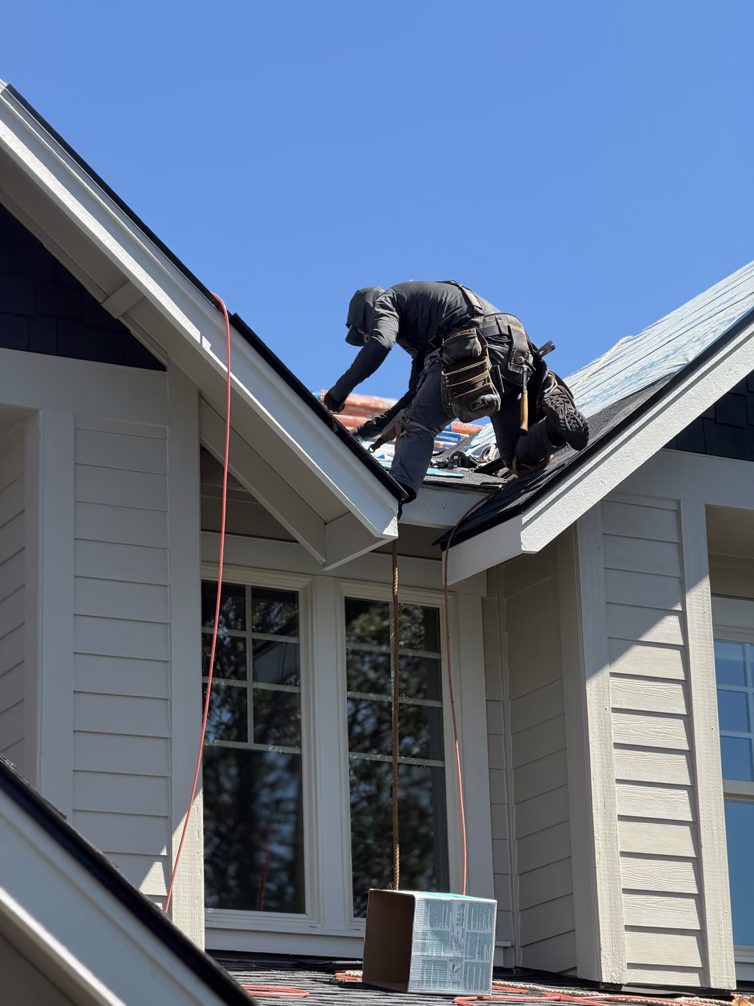 A construction worker wearing a tool belt is kneeling on a house roof installing or repairing roofing material under a clear blue sky, with a box suspended by a rope below near a window.