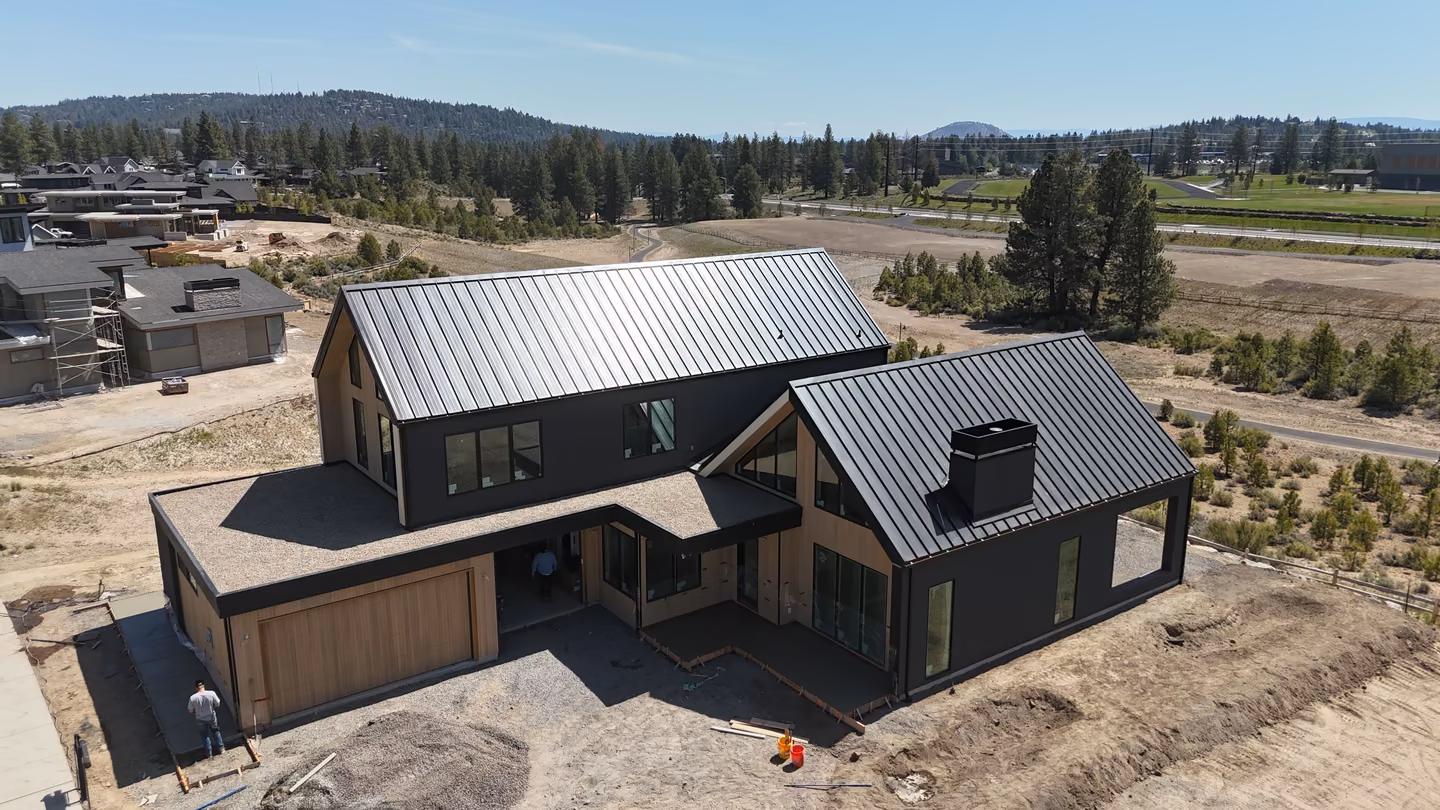 Aerial view of a modern two-story house under construction with dark exterior and metal roofs, surrounded by dirt and sparse trees in a developing neighborhood.