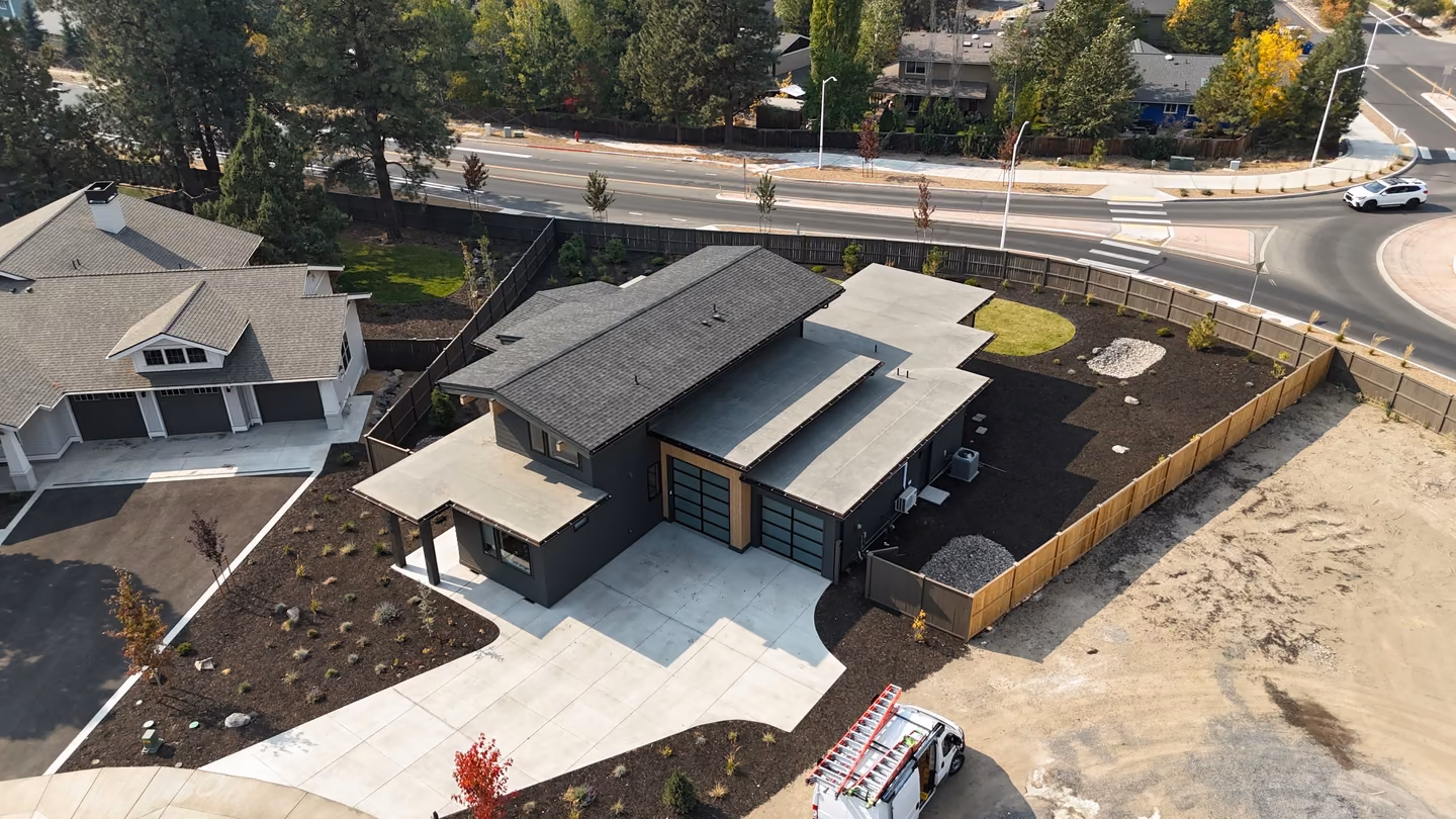A modern two-story house with dark exterior and flat and pitched roof sections, featuring a large concrete driveway, garage with frosted glass doors, and fenced backyard space, viewed from above in a suburban neighborhood.