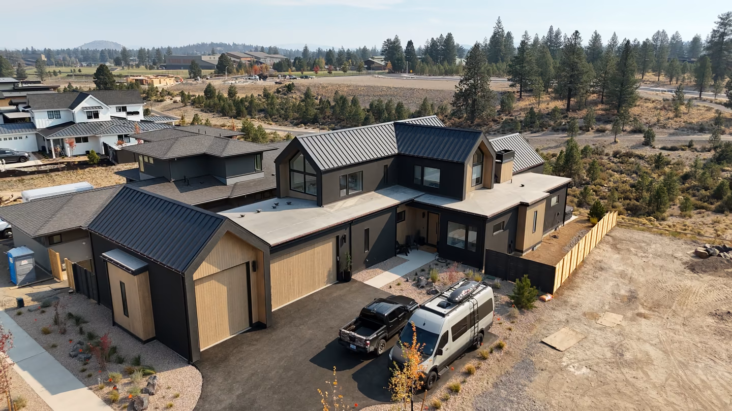 A modern, two-story house with black and natural wood exterior panels, flat and angled metal roofs, a three-car garage, and two vehicles parked in the driveway, surrounded by sparse landscaping and open land with trees in the background.