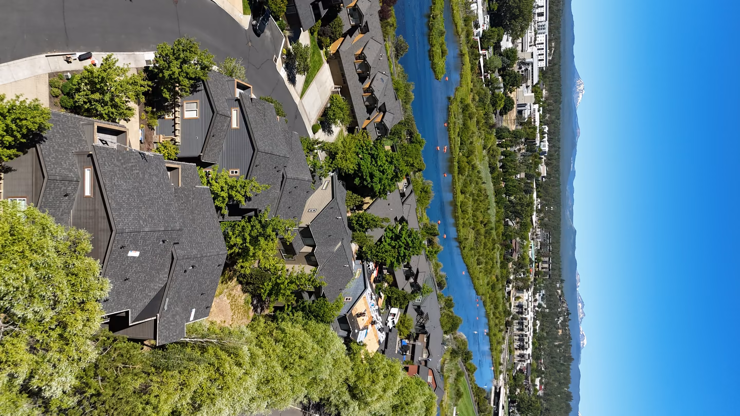 Aerial view of a residential neighborhood with rows of dark-roofed houses surrounded by green trees, a winding river with kayakers, and mountains with snow-capped peaks in the background under a clear blue sky.