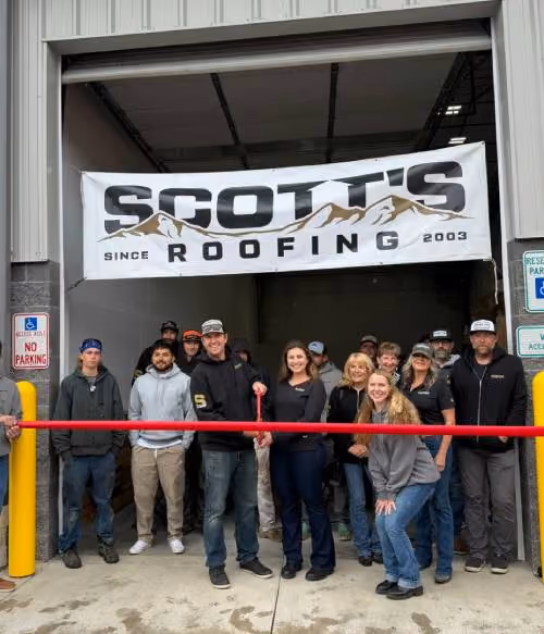 Group of people standing in front of a garage with a banner above reading 'Scott's Roofing Since 2003', cutting a red ribbon for a ribbon-cutting event.