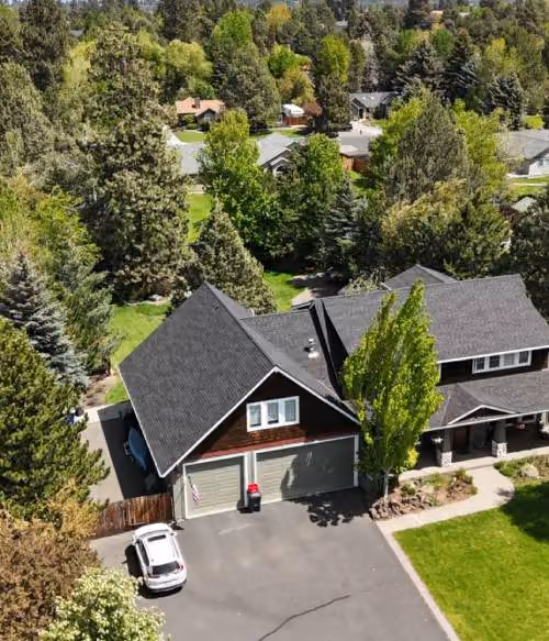 Aerial view of a residential neighborhood with houses surrounded by many green trees and a driveway with a white car parked near a house with a dark roof.