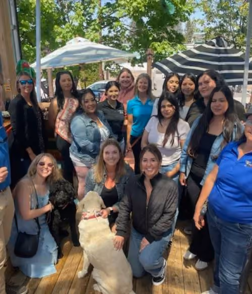 Group of fifteen diverse women gathered outdoors on a wooden deck under umbrellas, smiling and posing together, with two dogs in front.