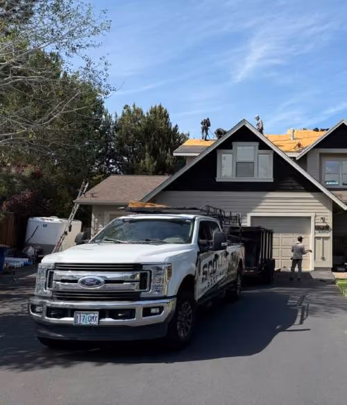 White pickup truck parked in the driveway of a two-story house with multiple workers installing or repairing the roof under a clear blue sky.