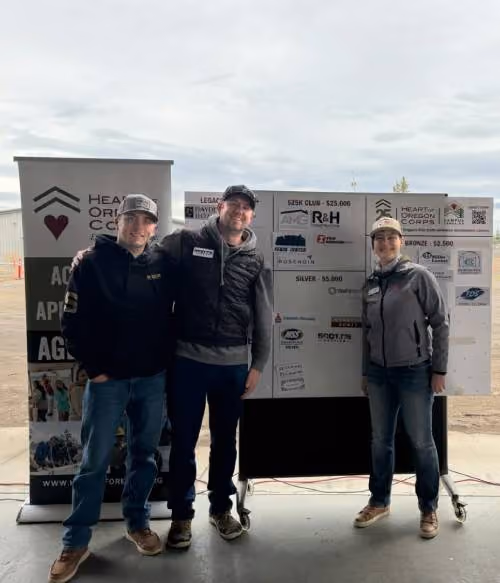 Three people standing in front of a banner and a signboard with sponsor logos at an event in an outdoor covered area. The banner has a heart logo and partially visible text, and the individuals are wearing casual outdoor clothing including hats and jackets.