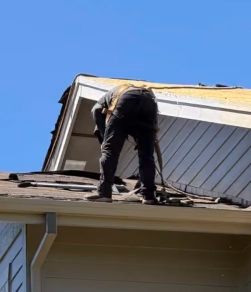 Worker wearing black pants and a safety harness replacing shingles on the roof of a house under a clear blue sky.