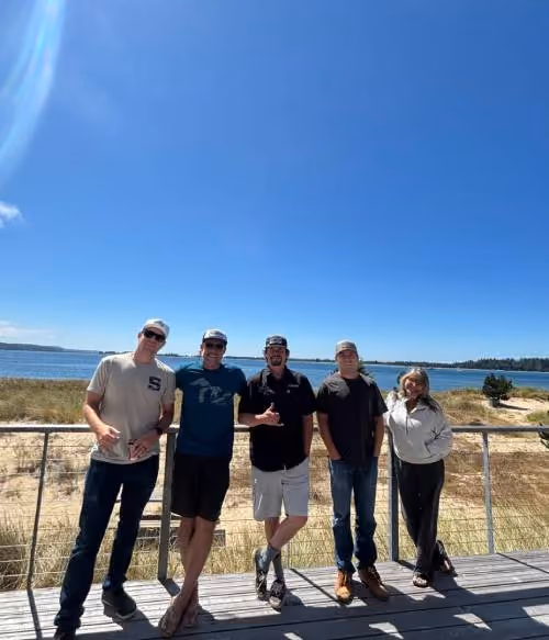 Five people standing on a wooden deck with metal railing in front of a scenic coastal backdrop with blue sky and water. Four men and one woman are casually dressed, enjoying the sunny day.