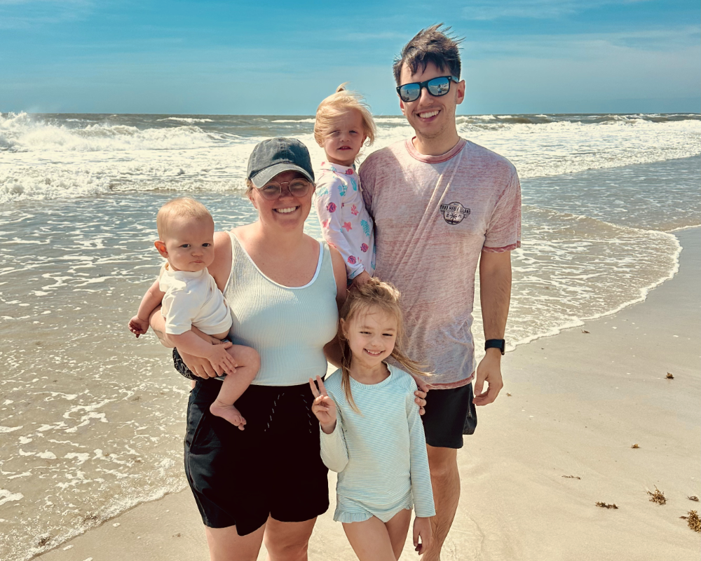 Smiling family of five posing on sandy beach with waves and blue sky in the background.