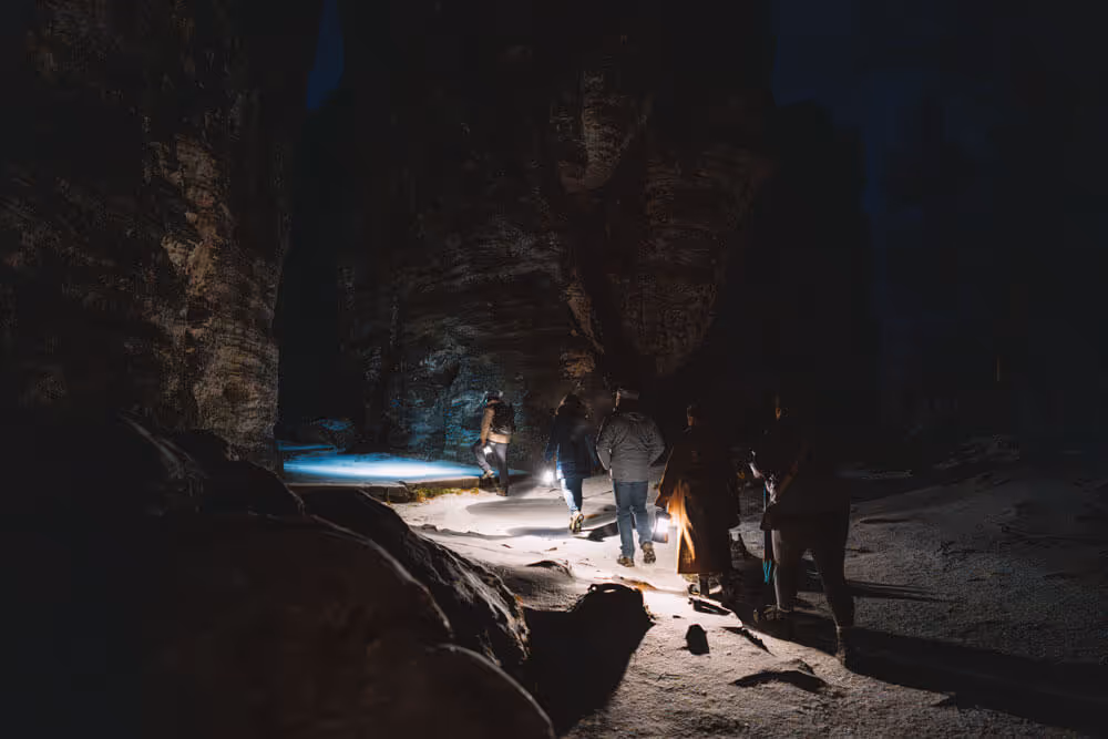Lantern-lit group walking between tall sandstone towers on a snowy trail at night at Tisa Rocks.