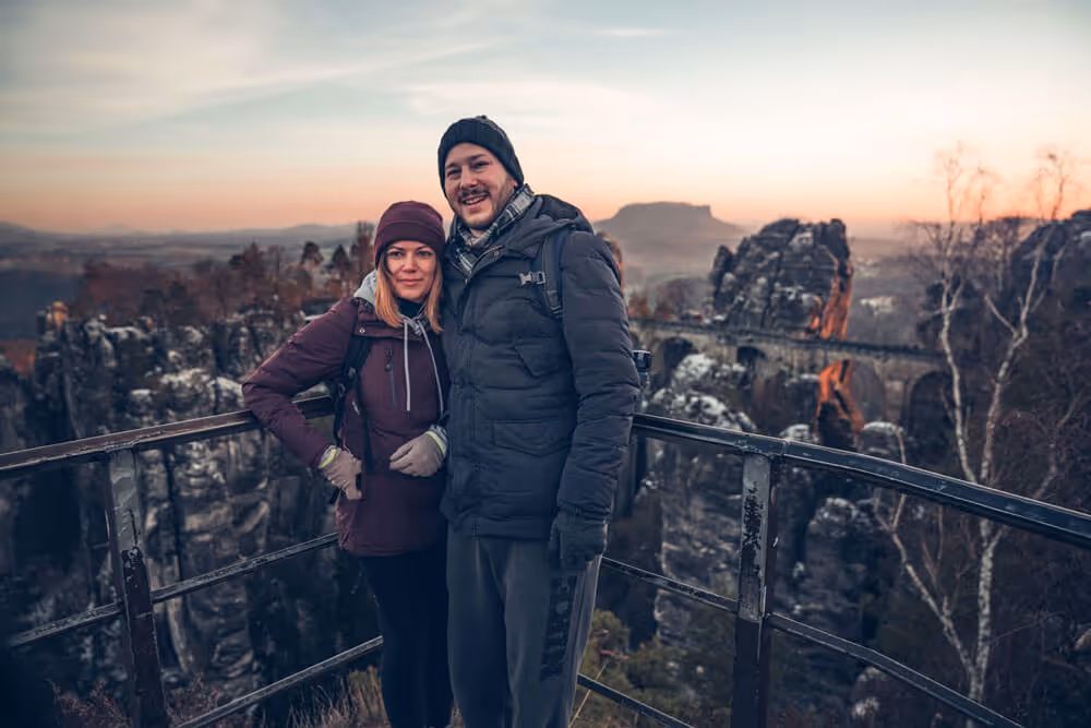 Couple posing on the snowy Bastei Bridge at sunrise, framed by golden light and sandstone cliffs.