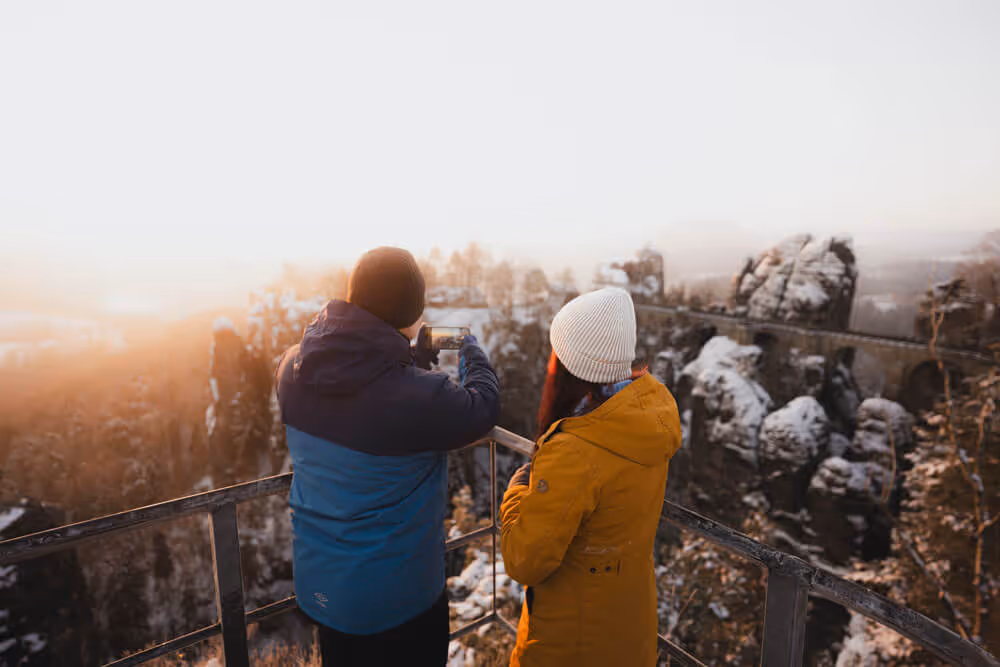 Two guests taking photos of the winter sunrise above Bastei Bridge and sandstone cliffs in Saxon Switzerland.