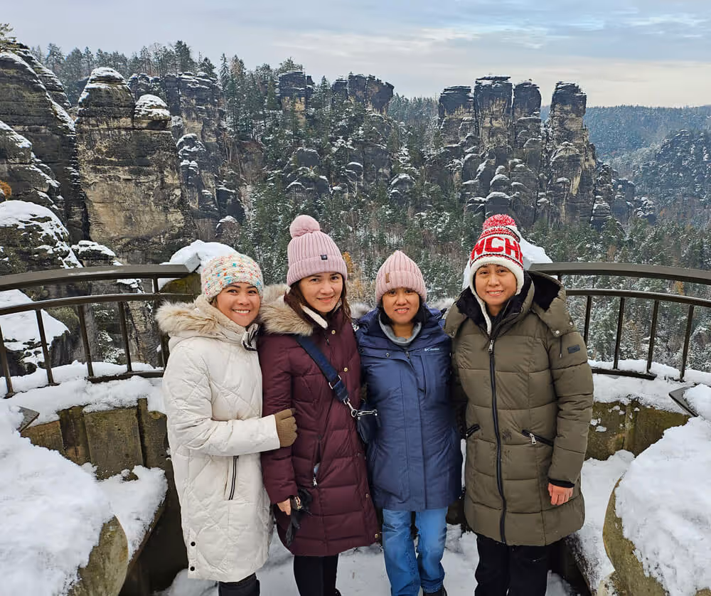 Four guests in winter coats posing at a snowy viewpoint with sandstone cliffs in Saxon Switzerland National Park.