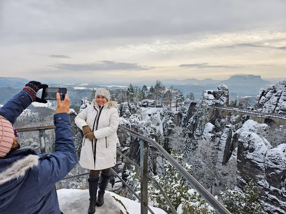 A smiling woman in a white coat posing for a photo with the snowy Bastei Bridge and cliffs behind her.