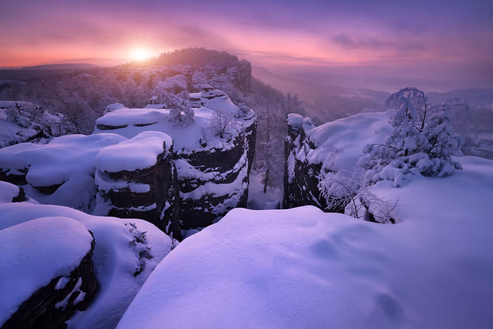 Snow-covered rock towers glowing in purple light at sunrise in Bohemian Switzerland National Park.