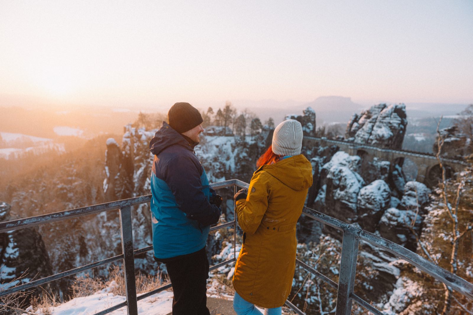 Two hikers at a viewpoint near Bastei Bridge above snowy sandstone rocks.