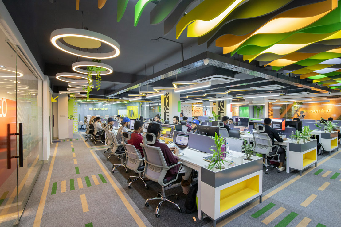 Modern office with employees working at desks arranged in rows under circular and wave ceiling lights with plants around.