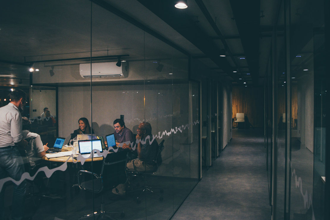 Group of five people collaborating around a conference table with laptops inside a glass-walled office room.