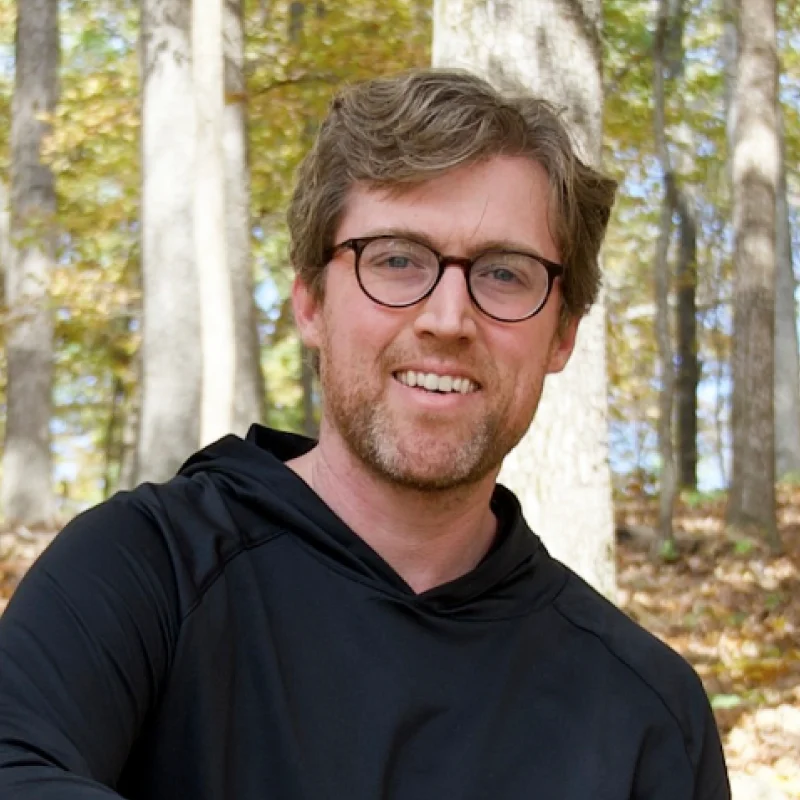 Smiling man with glasses wearing a black hoodie standing in a wooded area with autumn leaves.