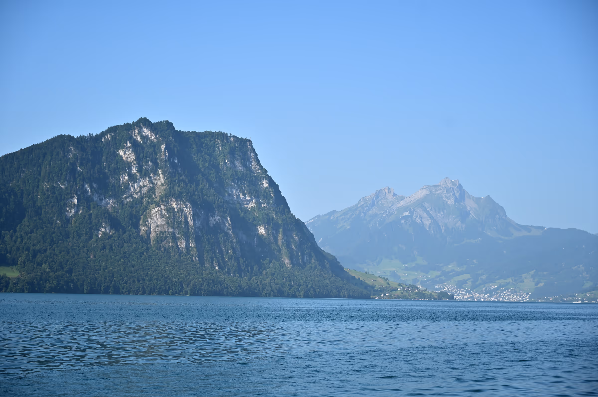 Calm lake water in the foreground with steep forested cliffs on the left and distant alpine peaks under a clear blue sky.