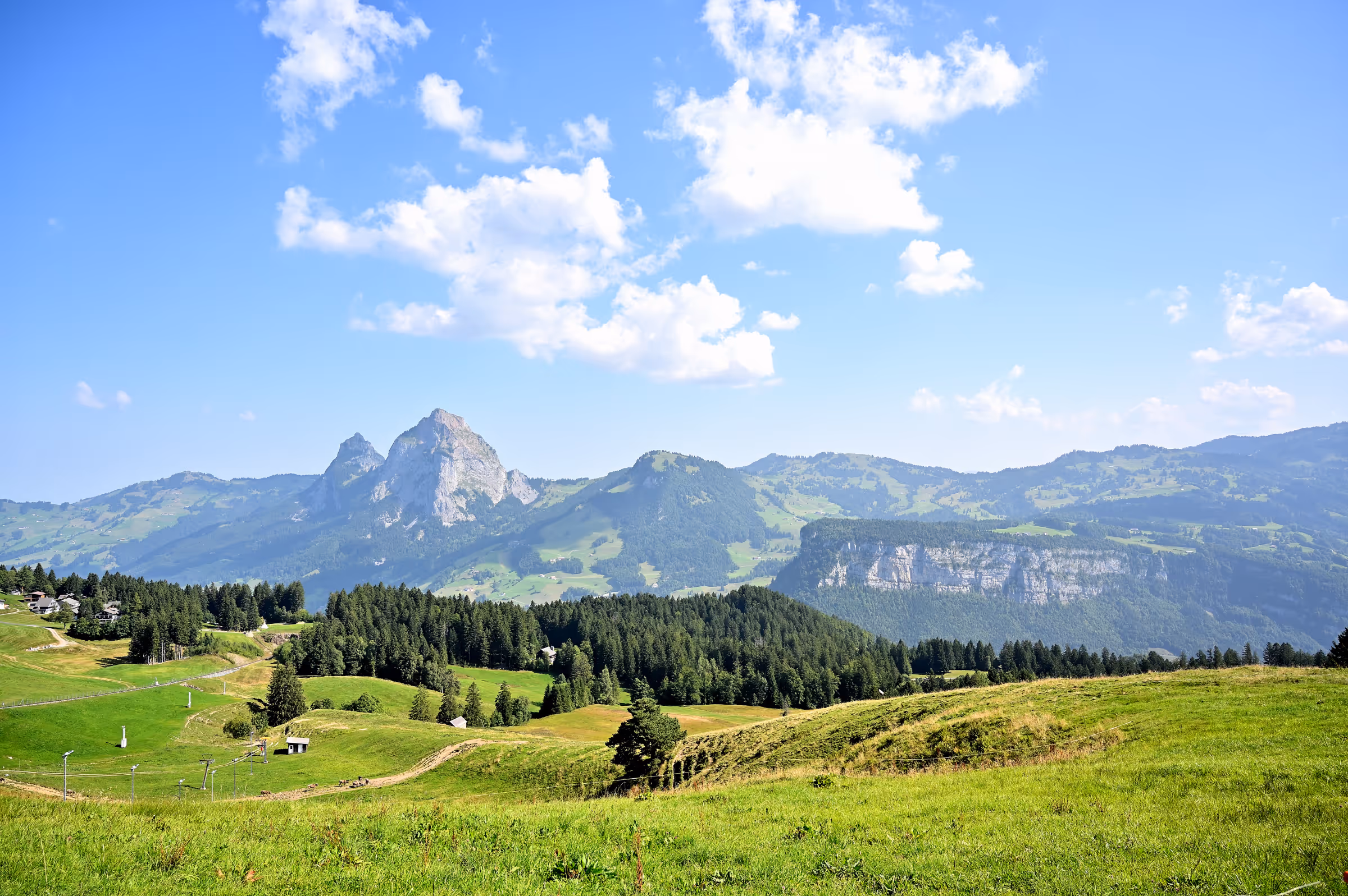 Rolling alpine meadows near the Stoos Ridge with forested hills and rugged mountain peaks beneath a blue sky with scattered clouds, Switzerland.