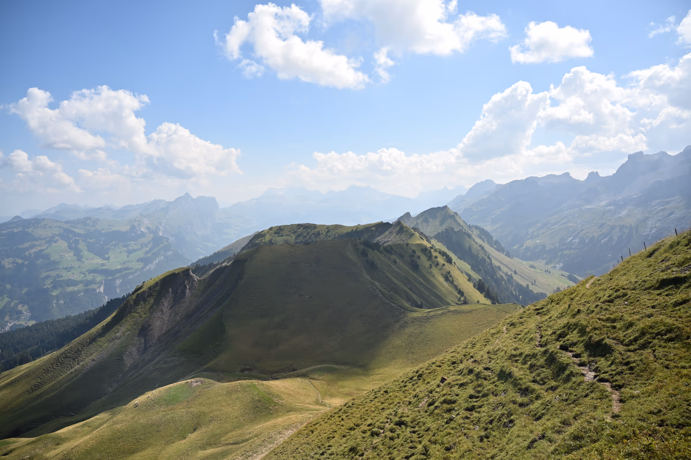 Panoramic view from a grassy alpine ridgeline with rolling green peaks and layered mountain ranges under a partly cloudy sky, near Stoos Ridge, Switzerland.