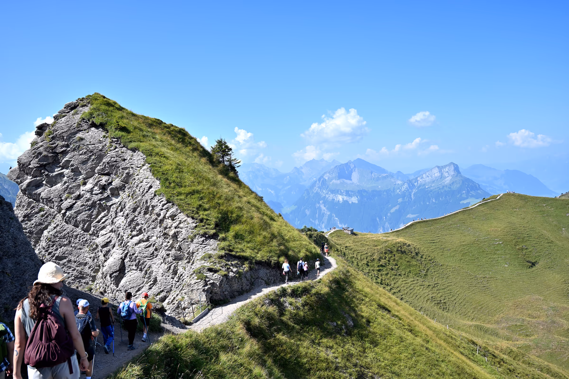 Hikers walking along a narrow alpine trail carved into a grassy ridgeline with steep rock faces and distant mountain peaks under a clear blue sky, near Stoos Ridge, Switzerland.