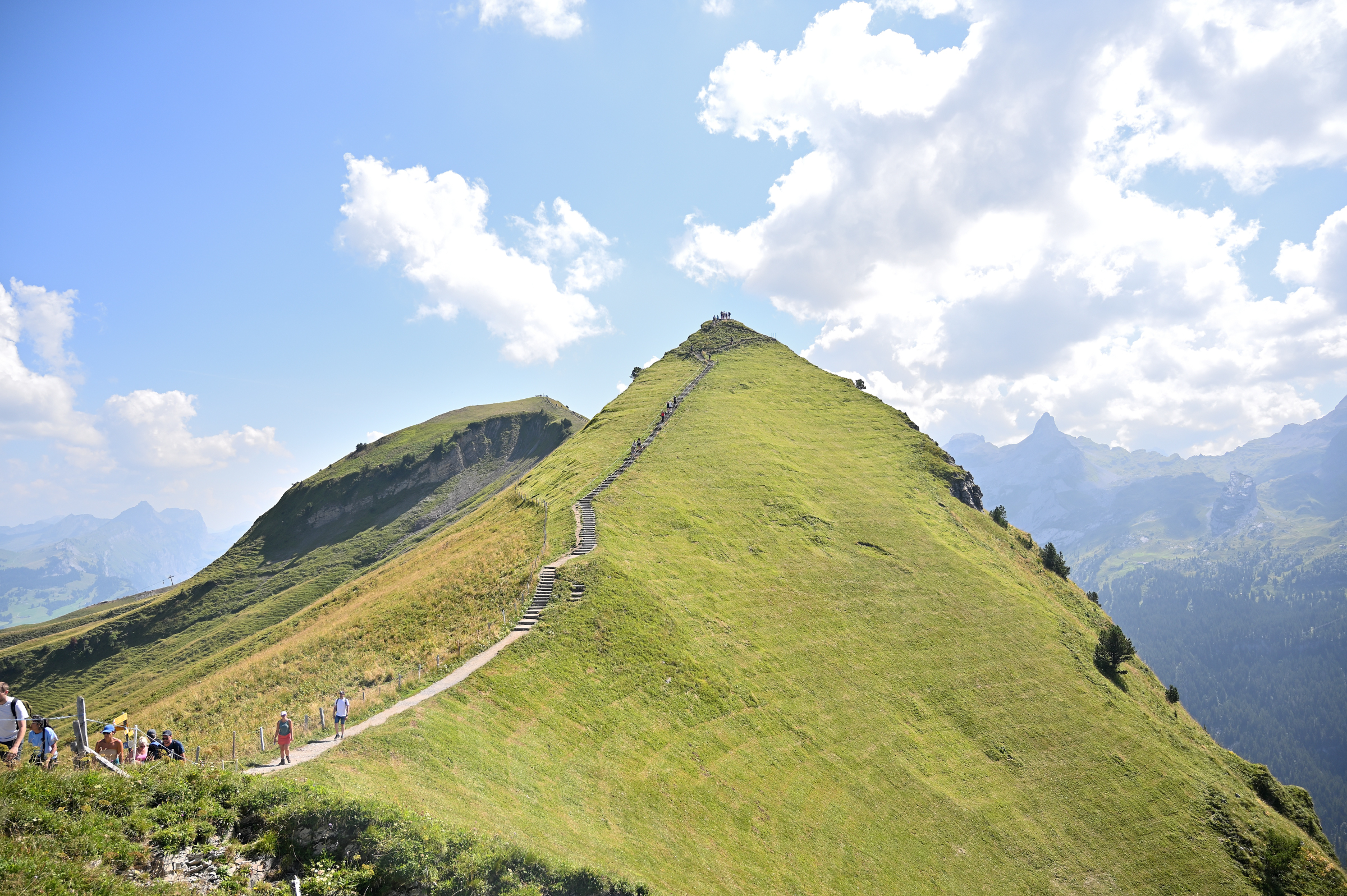 Hikers ascending a grassy alpine ridgeline toward a prominent peak, with steep slopes and distant mountain ranges under a clear blue sky near Stoos Ridge, Switzerland.