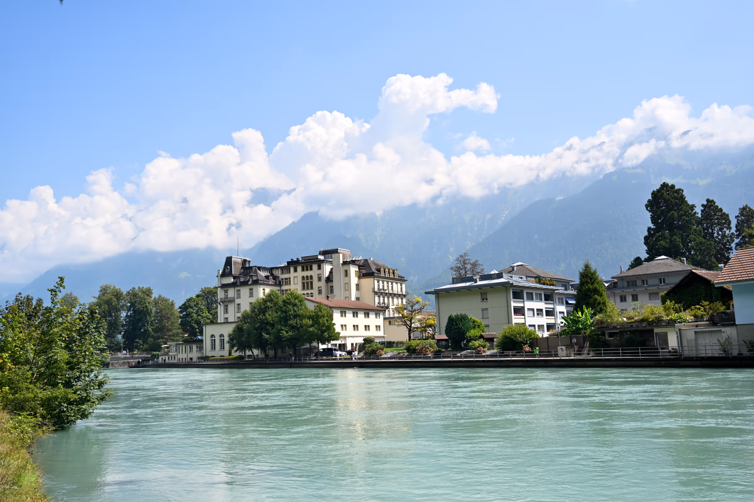 Clear turquoise water of the Aare River flowing past riverside buildings and trees in Interlaken, Switzerland, under a bright summer sky.