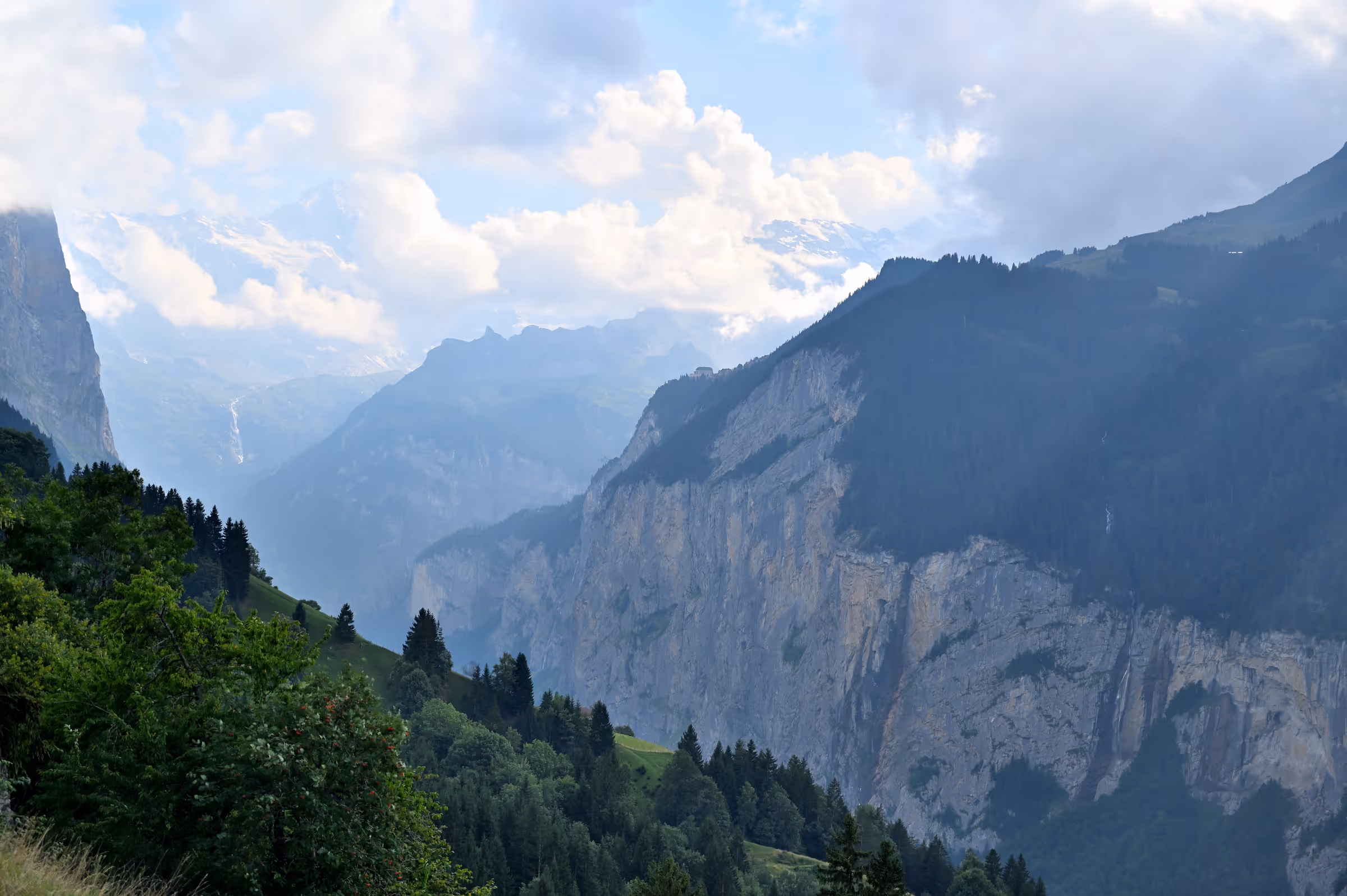 View over the Lauterbrunnen Valley from Wengen, with steep alpine cliffs, forested slopes, and layered mountain peaks under a partly cloudy sky in Switzerland.