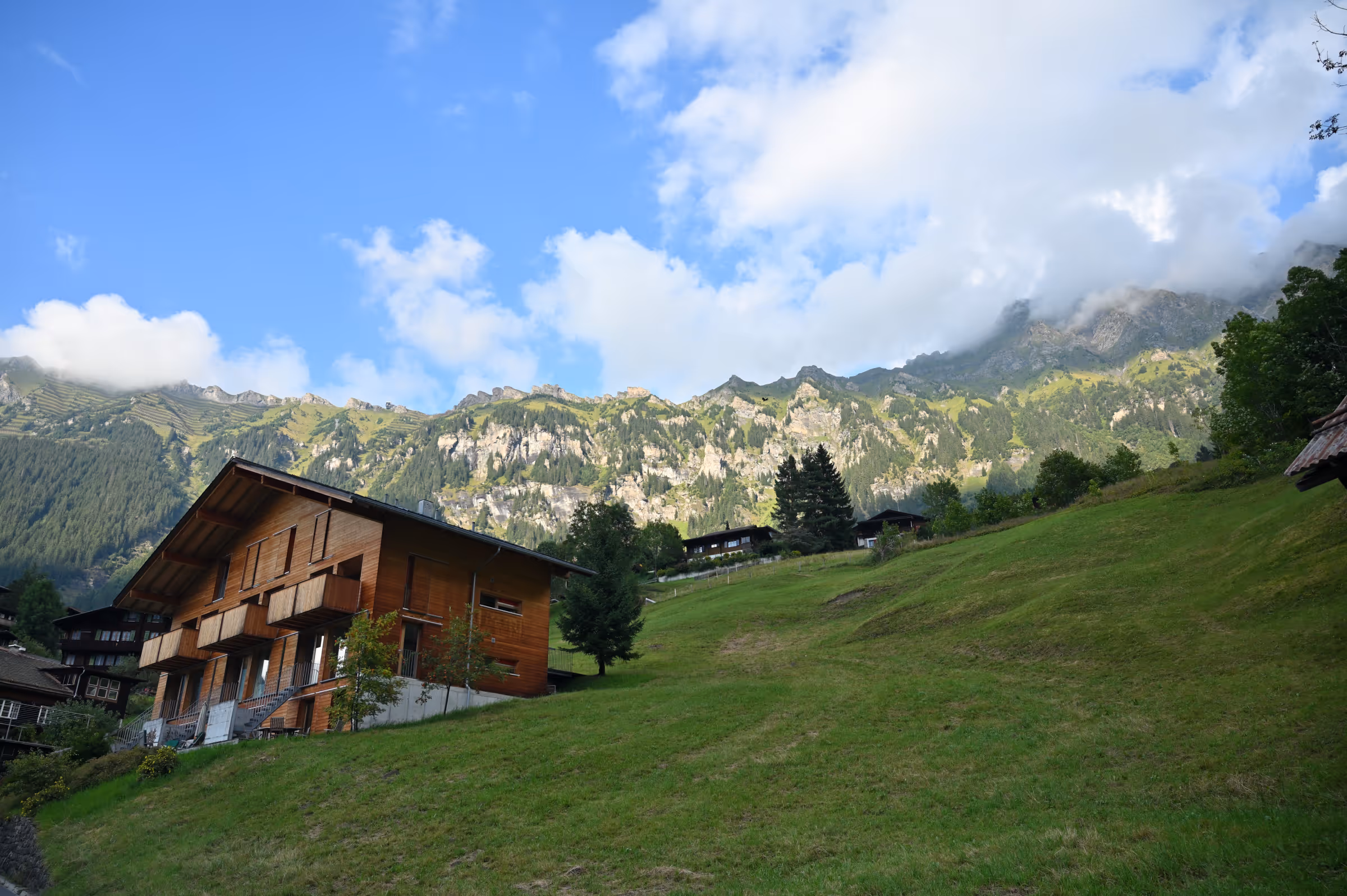 A wooden alpine chalet on a grassy hillside with mountain slopes and scattered homes rising above the village of Wengen, Switzerland, under a partly cloudy sky.