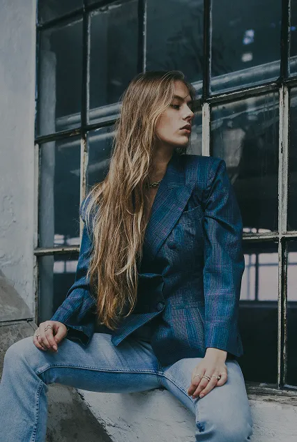 Young woman with long hair wearing a blue plaid blazer and jeans sitting by a large industrial window.