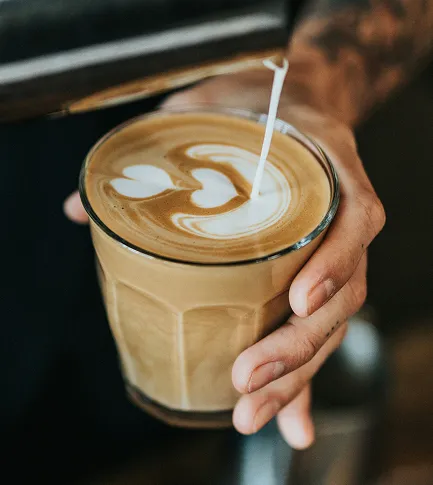 Hand holding a glass of latte with heart-shaped latte art as milk is being poured.