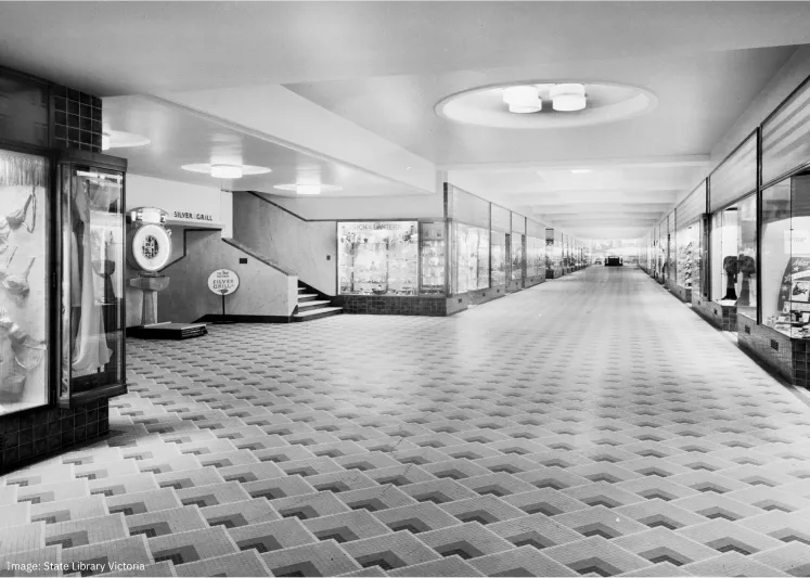 Black and white photo of a long, empty shopping arcade with tiled zigzag flooring, display windows on both sides, ceiling lights, and a sign for Silver Grill near a staircase.