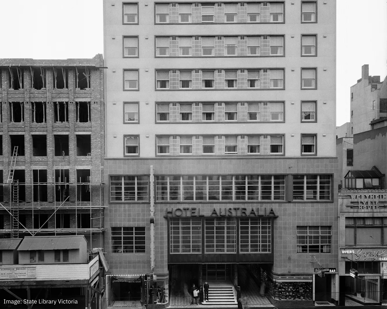 Black and white photo of the Hotel Australia building with a tiled facade and multiple windows, an adjoining under-construction brick building on the left, and several people standing near the entrance.