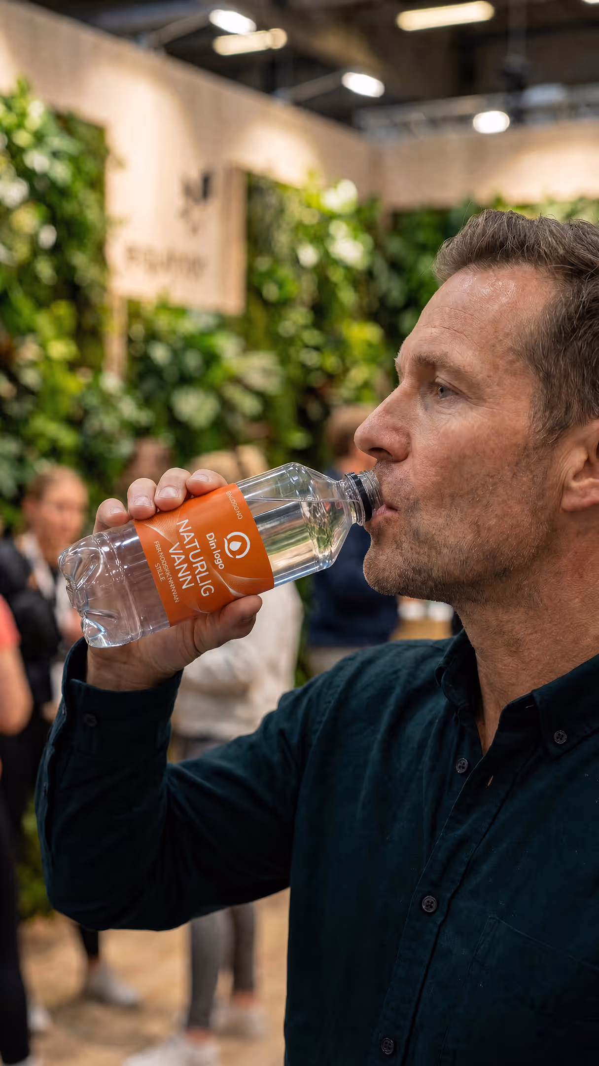 Mann drikker vann fra en plastflaske med oransje etikett i et innendørs grønt miljø.