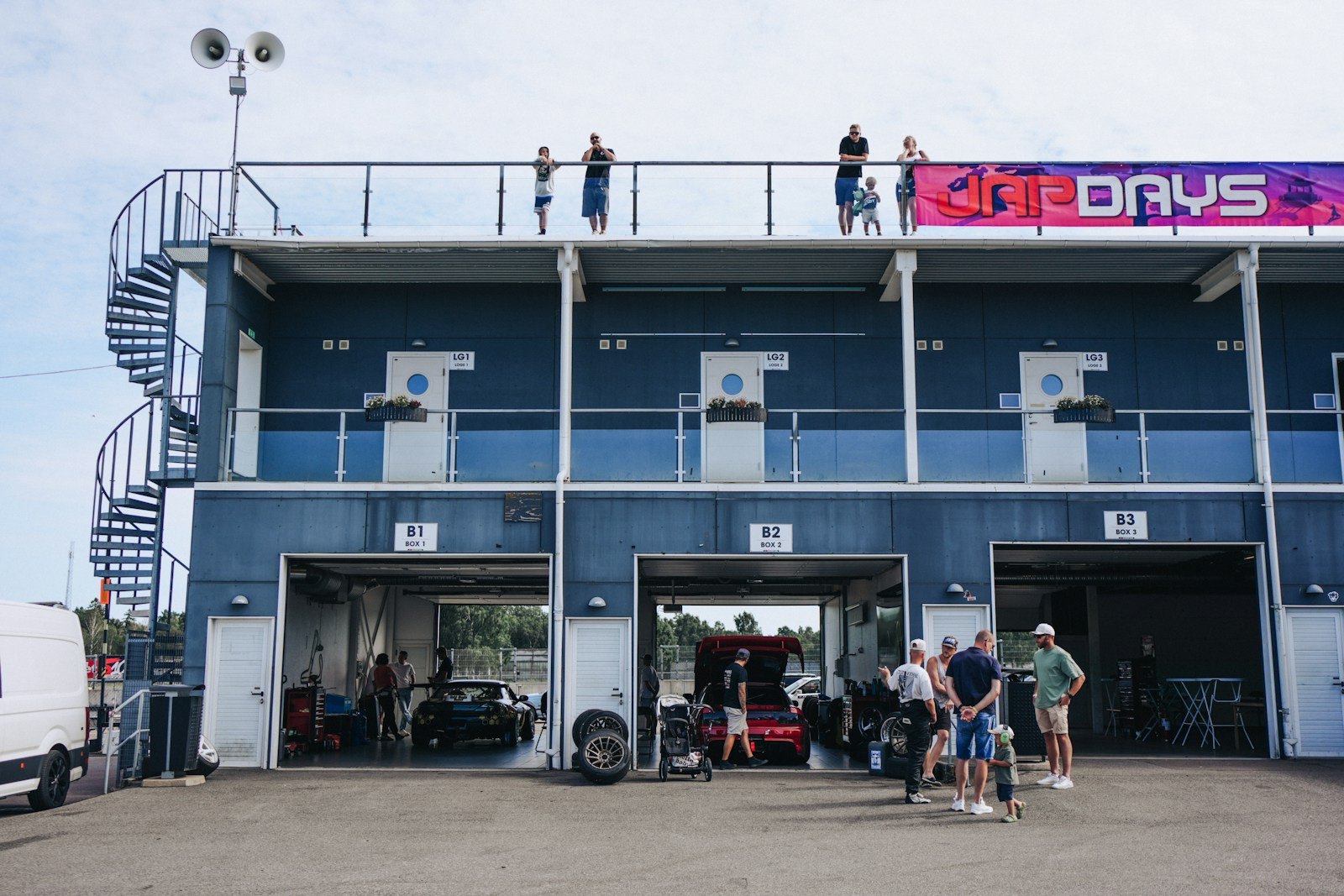 Des spectateurs sur la terrasse au-dessus des stands d'un circuit automobile, les garages ouverts en dessous