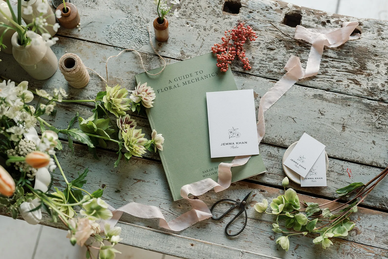 Rustic wooden table with green floral book, gardening shears, beige ribbon, business cards, and assorted fresh flowers.
