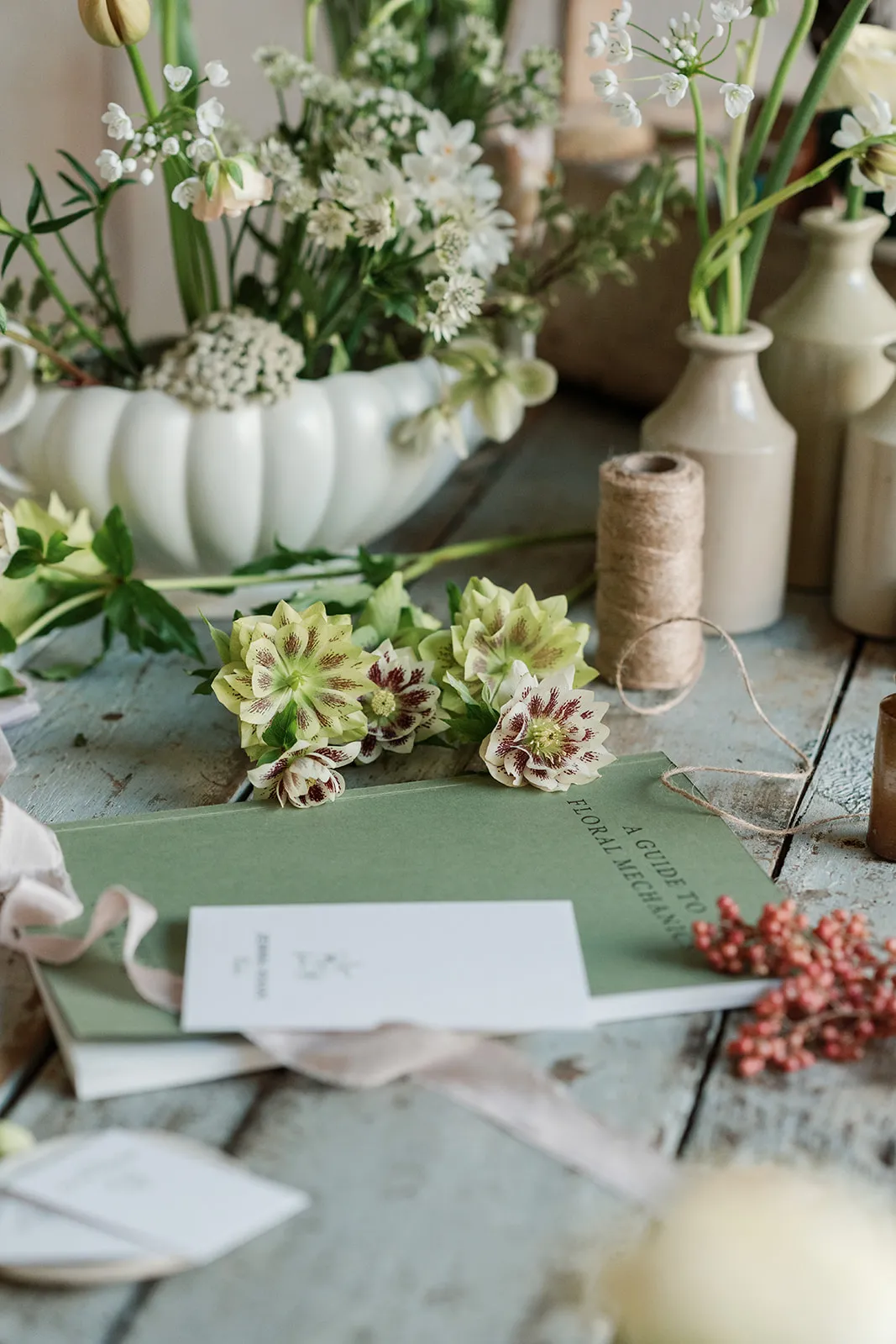 A close-up of a green book titled 'A Guide to Floral Mechanics' surrounded by cream and burgundy flowers, beige ceramic vases, twine, and a white card on a rustic wooden table.