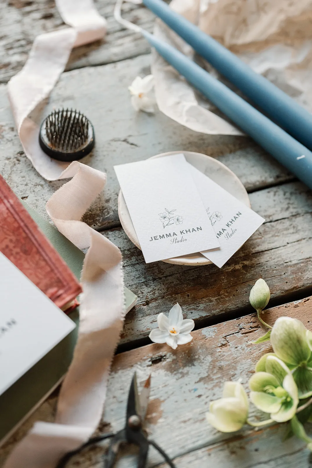 Rustic wooden table with Jemma Khan Studio business cards, blue candles, scissors, ribbon, and green flowers.