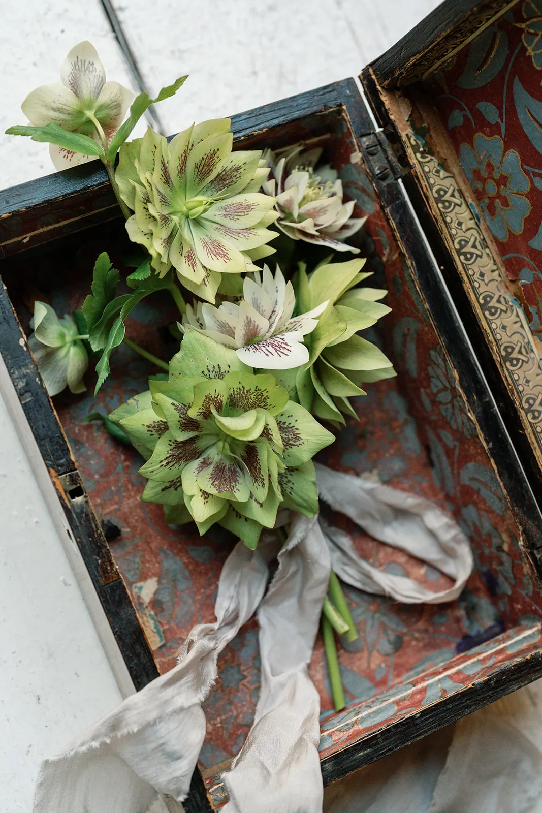 Light green and white flowers with dark speckles arranged inside an open vintage wooden box lined with patterned fabric, alongside a frayed white ribbon.