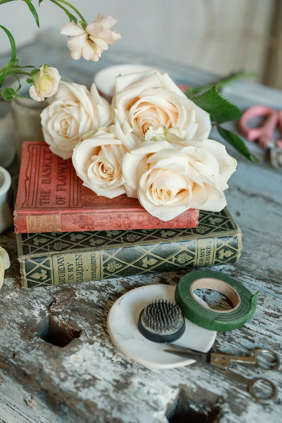 Four cream-colored roses resting on two vintage books on a rustic wooden table with floral arranging tools nearby.