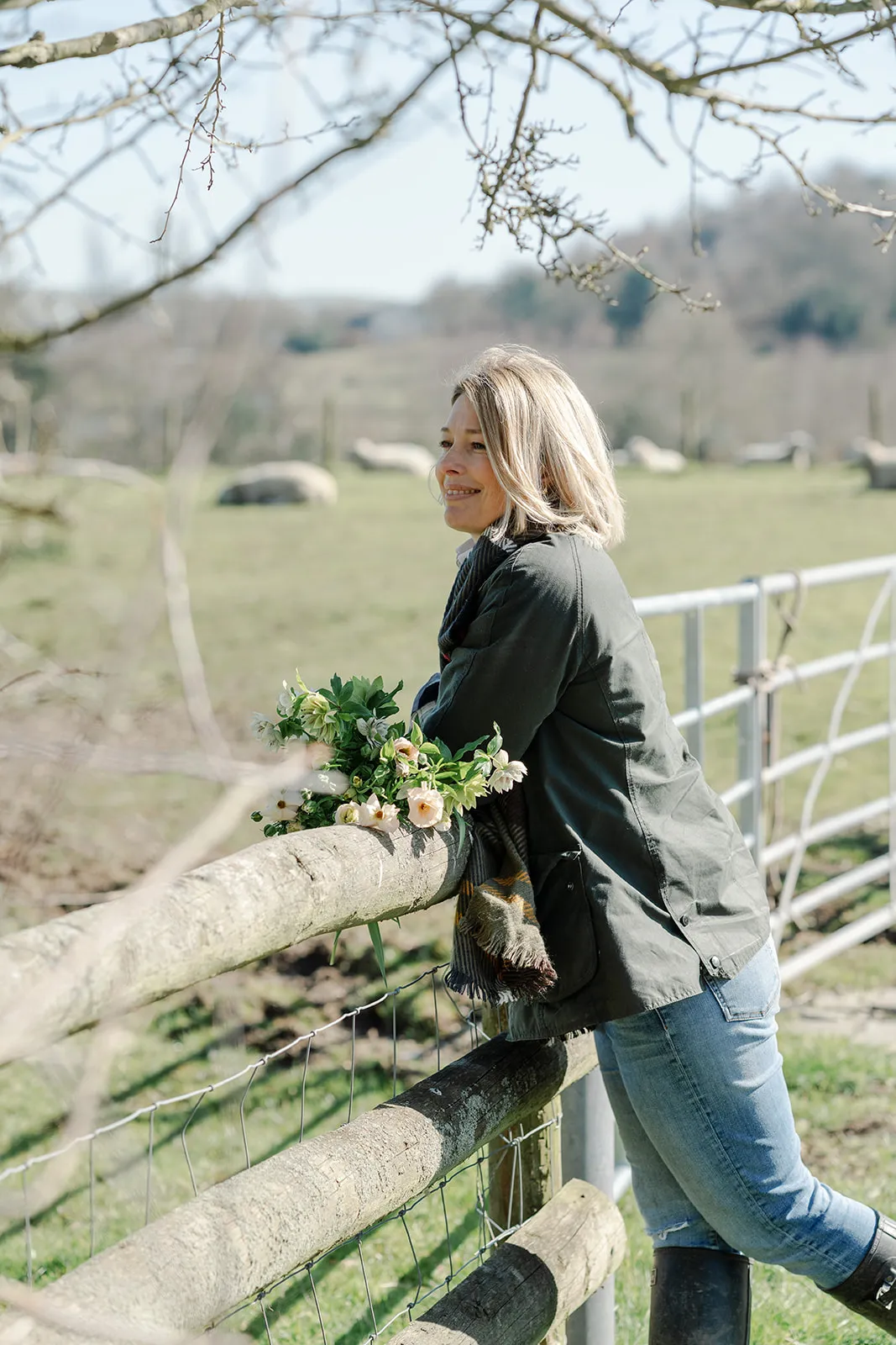 Smiling woman leaning on a wooden fence with a bouquet of flowers in a grassy field with sheep in the background.