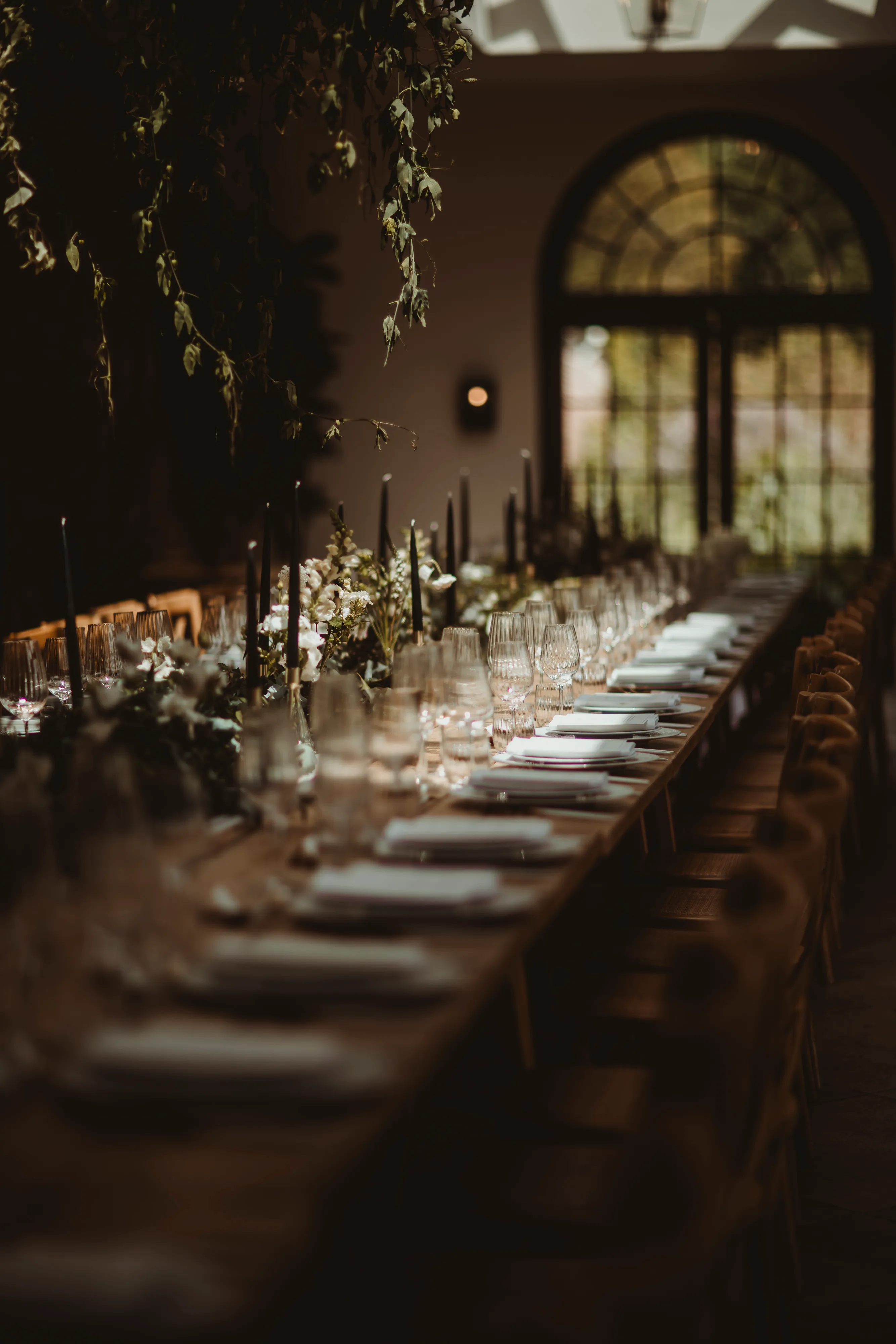 Long dining table set with glassware, plates, black candles, and floral centerpieces in a dimly lit room with arched windows.