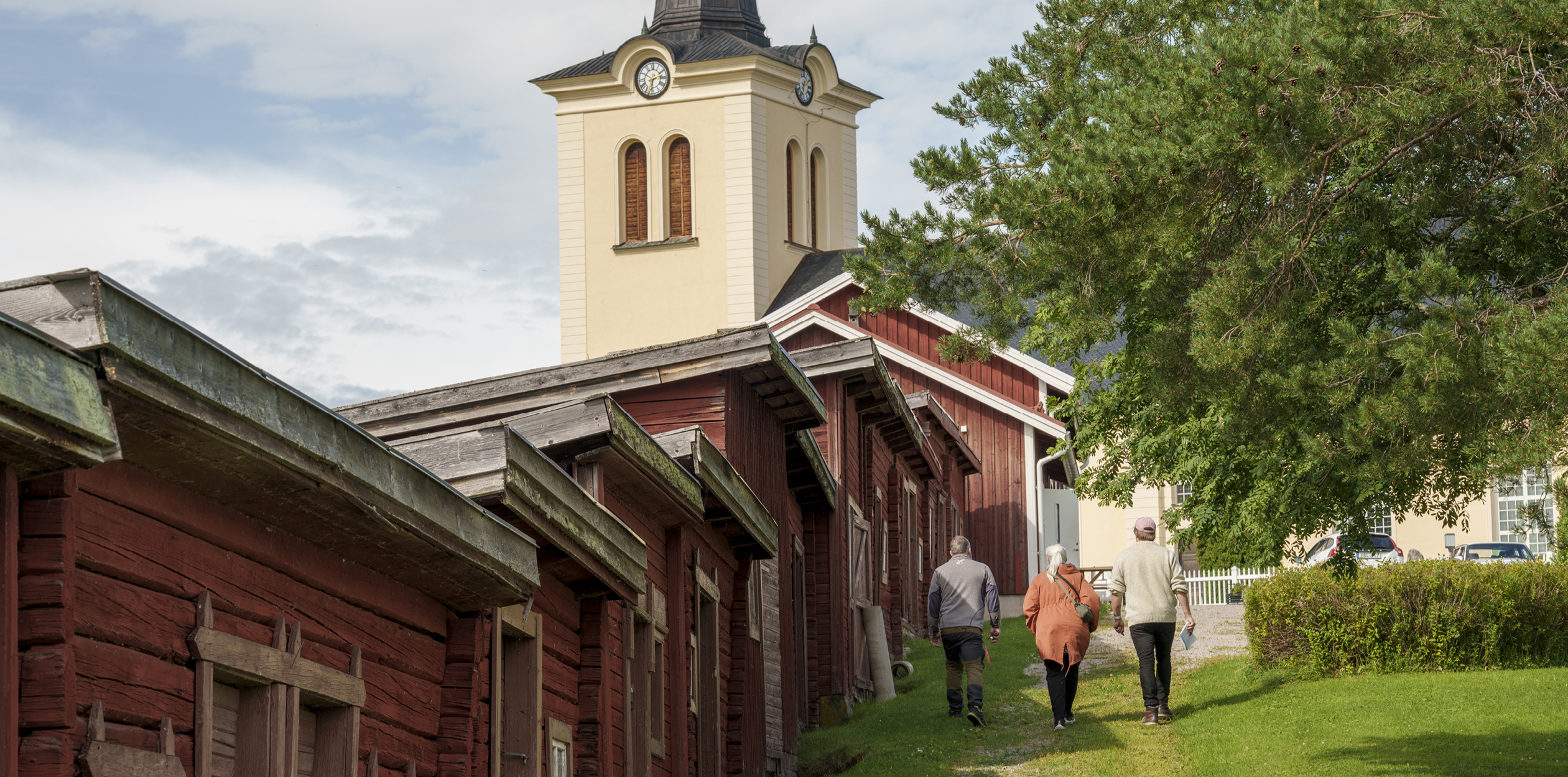 Gamla röda timrade byggnader i Revsund, med kyrkan i bakgrunden. Tommy, Malin och Linus går bredvid byggnaderna. 