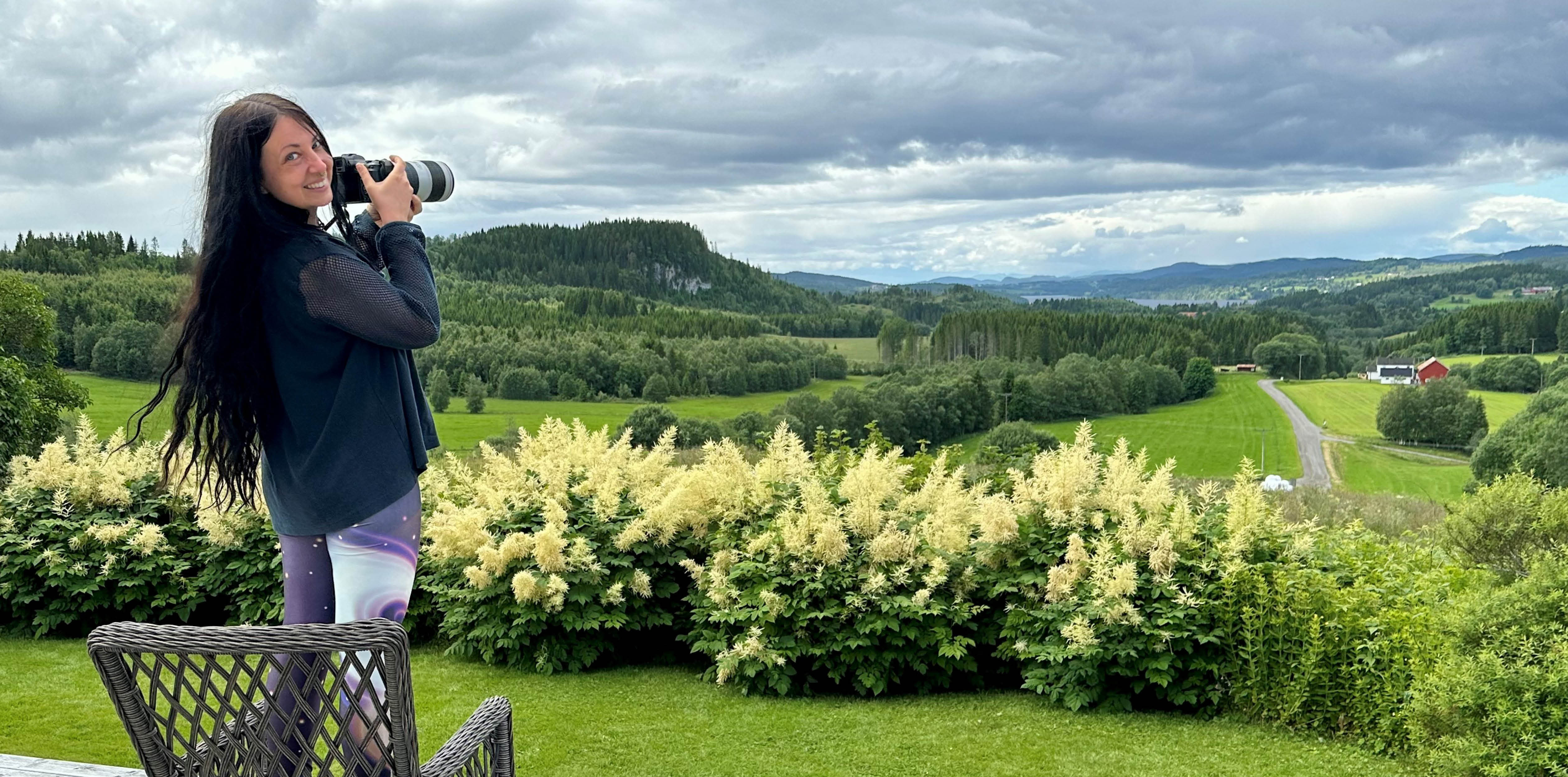 Cecilia Kreuzberger står utomhus på Troset Gård med utsikt över gröna fält, skog, och böljande kullar. Hon håller i en egen kamera och tittar leende in i fotografens kamera. 