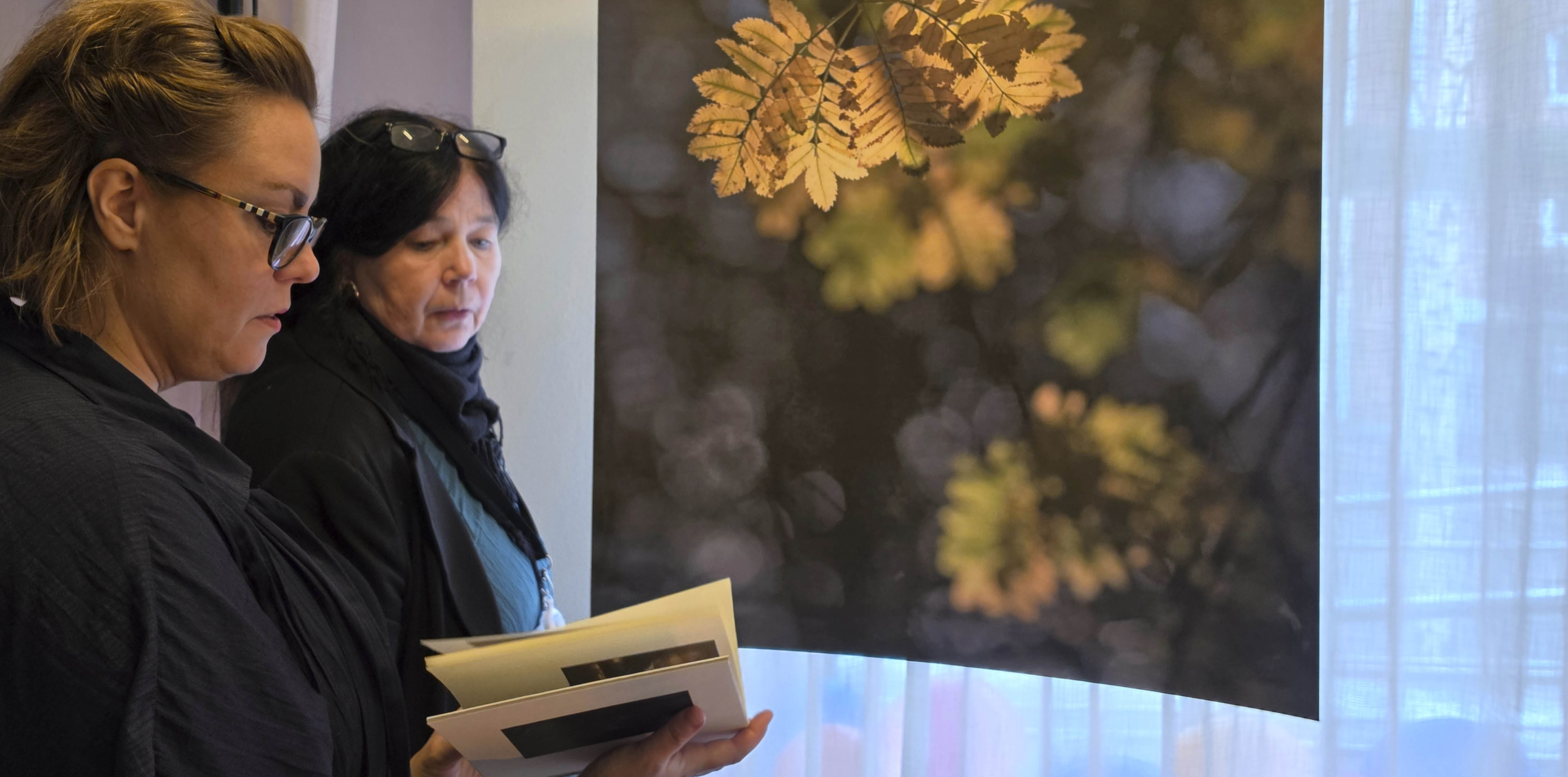 Anna Vidmark bläddrar i boken Stien til Troset och visar för Eva Aspling. I bakgrunden hänger ett stort fotografi med höstfärgade löv. 