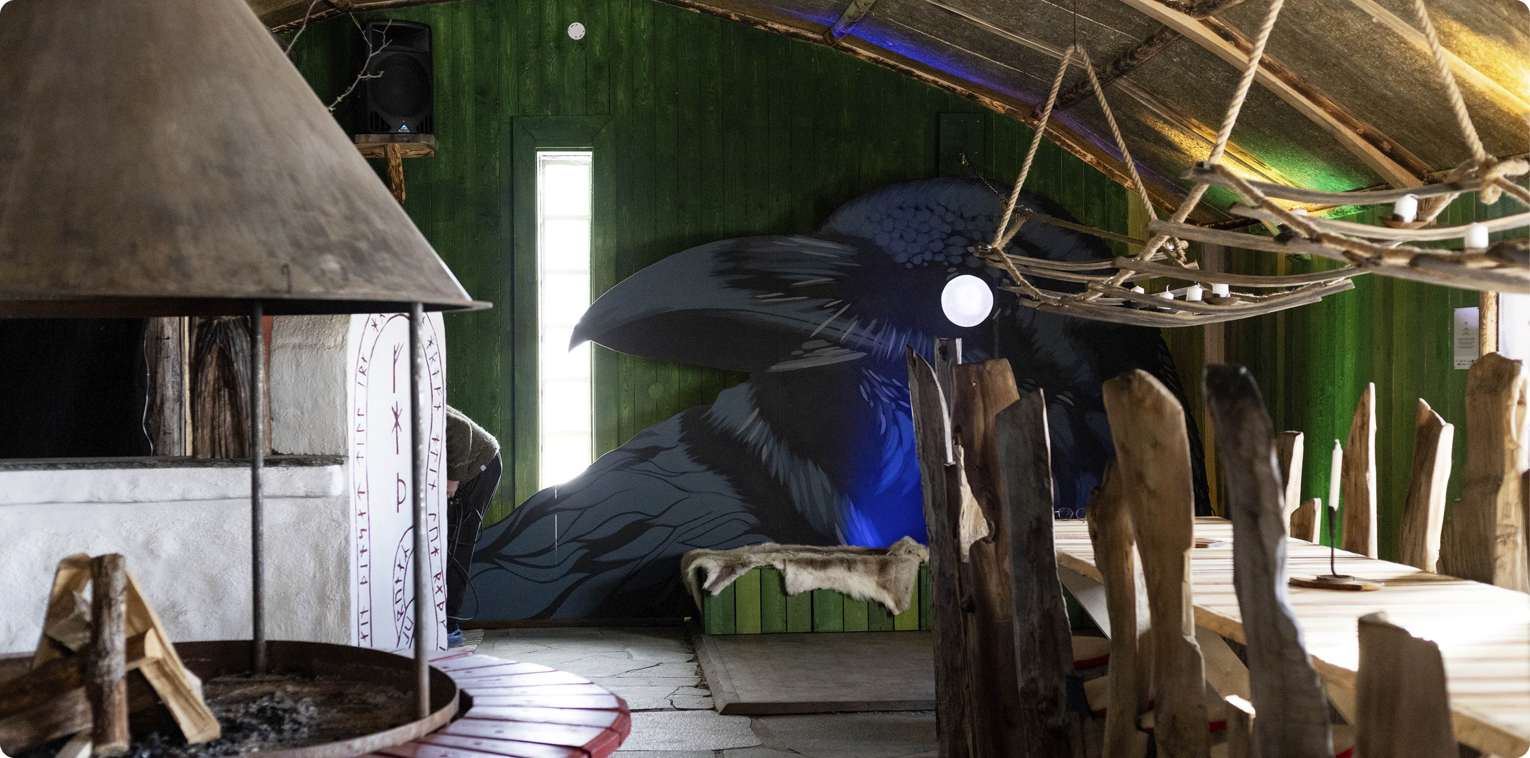 Miljöbild från huset Ögat. I förgrunden en öppen eldplats med ved till vänster, och specialtillverkade stolar och bord i trä  till höger. mot den bakre väggen syns en stor målning av en korp. 