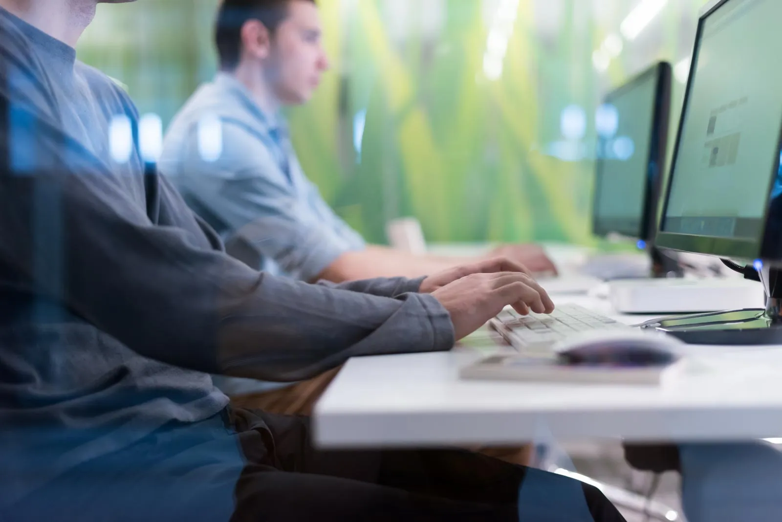 Two men typing on keyboards at a row of desktop computers in an office setting with a green background.