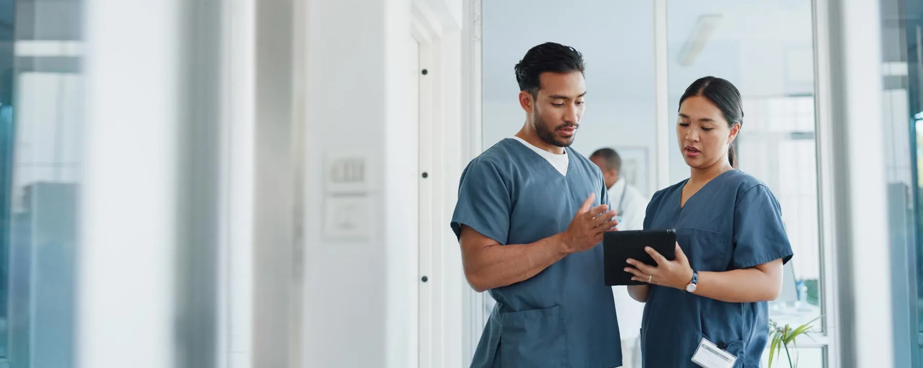 Two healthcare professionals in scrubs discussing information on a digital tablet in a hospital hallway.